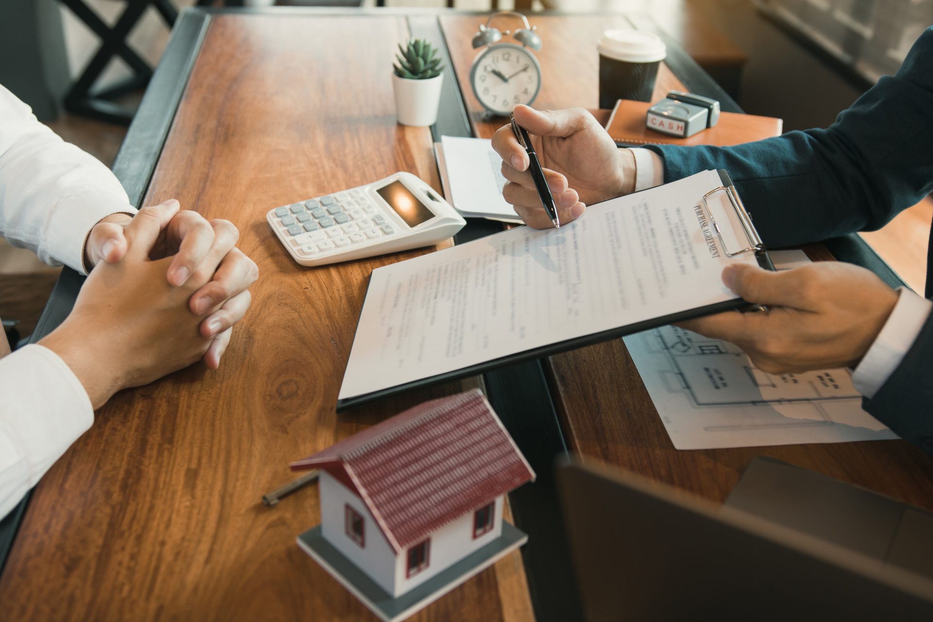 man holding a home agreement form, gesturing with a pen to another man with folded hands, representing mortgage process